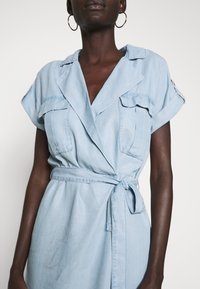 Jasnoniebieska sukienka z denimowej tkaniny, o kroju kopertowym, z krótkimi rękawami, z dwiema kieszeniami na piersi i paskiem do wiązania w talii. Gładka tekstura, casualowy design.