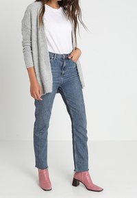 Cardigan grigio a maglia sopra una t-shirt bianca, abbinato a jeans blu a vita alta e stivaletti rosa con tacco largo.