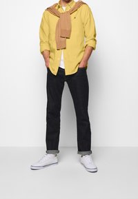 Gele blouse met knopen, wit ondershirt, bruine trui om de nek, donkerblauwe spijkerbroek en witte sneakers met blauwe accenten.