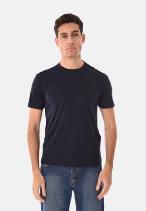 Uomo in piedi, rivolto in avanti, che indossa una maglietta a maniche corte blu scuro semplice e jeans blu su sfondo bianco.