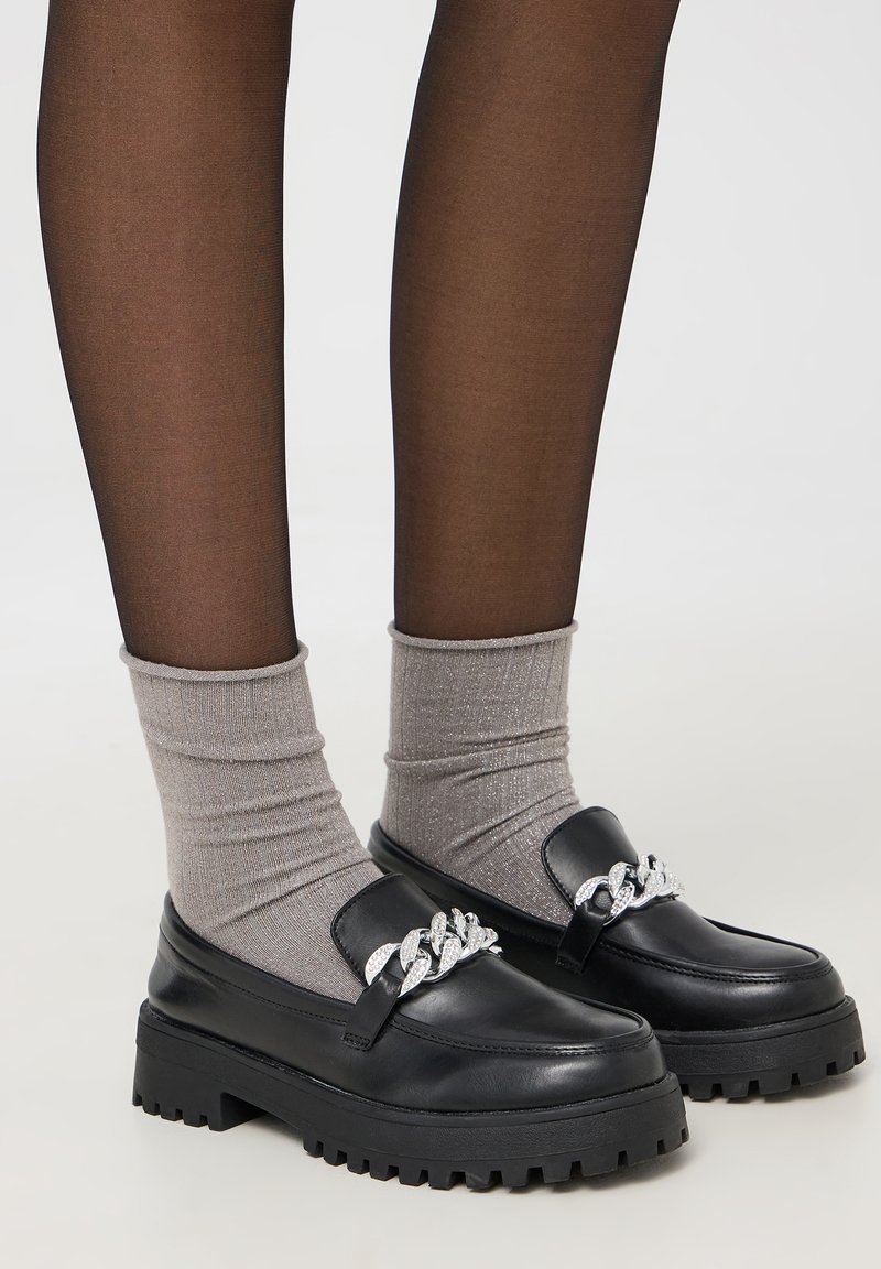 Mocassini in pelle nera con suola spessa, adornati da un accento decorativo di catena argentata. Indossati con calzini a coste grigi.