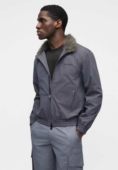 Γκρι ζακέτα με φερμουάρ, με ψεύτικη γούνα στον γιακά, ελαστικές μανσέτες και τελείωμα. Φορεμένη πάνω από ένα μαύρο πουκάμισο, διαθέτει λογότυπο στο αριστερό στήθος.