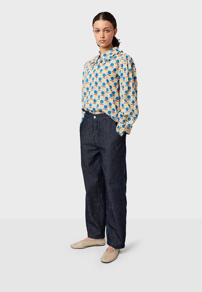 Bedruckte Bluse mit blauen und roten Kreisen auf einem cremefarbenen Hintergrund, kombiniert mit marineblauen gestreiften weit geschnittenen Hosen und beigen flachen Schuhen.