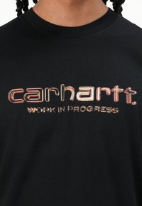 Black cotton t-shirt featuring a colorful, shiny "carhartt." logo and "WORK IN PROGRESS" text across the chest in a contrasting design.
