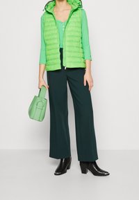 Gilet imbottito verde con chiusura a zip, abbinato a un top a maniche lunghe coordinato e pantaloni larghi verde scuro. Stivali neri con tacco e borsa verde.