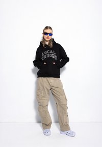 Czarna bluza z kapturem z napisem "LOCAL HEROES", beżowe spodnie cargo z kieszeniami oraz białe klapki. Model nosi niebieskie okulary przeciwsłoneczne.