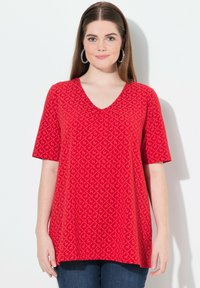 Camiseta roja de cuello en V con mangas cortas, tela texturizada que presenta un patrón de diamantes, con aberturas laterales y un corte holgado. Llevada con jeans de color azul oscuro.