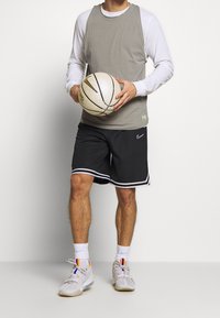 Muž v šedém bezrukávovém tričku přes bílé dlouhé rukávy, v černých basketbalových šortkách, držící bílé basketbalové míče Nike na bílém pozadí.