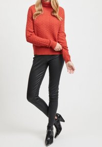 Roter kabelgestrickter Rollkragenpullover kombiniert mit schwarzen, taillierten Lederhosen und schwarzen, hochhackigen Stiefeletten, die strukturierte Materialien und ein elegantes Design präsentieren.