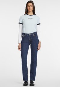 Camiseta de color azul claro con ribete azul oscuro, que presenta el texto "GUESS JEANS"; combinada con pantalones vaqueros de tiro alto en azul oscuro y zapatos negros.