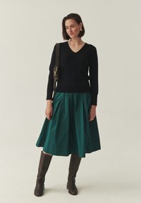 Pull noir à col en V, jupe plissée turquoise, bottes en cuir marron montant jusqu'aux genoux. Le mannequin porte un sac à main à bandoulière marron. Design simple et épuré.