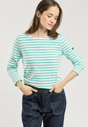 Femme portant une chemise blanche à manches longues avec des rayures horizontales vert clair et un pantalon en jean bleu foncé, les mains dans les poches.