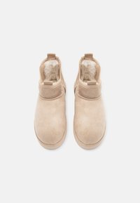 Bottes de cheville en suède beige avec une doublure douce et duveteuse, des orteils arrondis et des languettes robustes et courtes à l'arrière pour un enfilage facile.