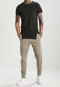 Schwarzes Baumwoll-T-Shirt mit einer Tasche und abgetragenen Details, kombiniert mit hellgrauen Sweatpants, die Patches und gerippten Bündchen aufweisen. Weiße Sneakers.
