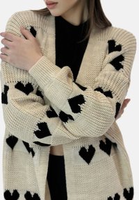 Stickad cardigan i krämfärg med svarta hjärtmönster, med en lös passform, långa ärmar och ribbade muddar. Texturen är grov och mysig.