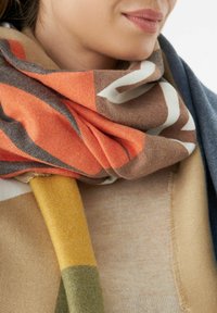 Femme portant une écharpe multicolore avec des motifs orange, marron, jaune et vert enveloppée autour de son cou et une veste bleue.