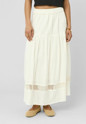 Femme portant une jupe blanche longueur cheville avec un motif ajouré et des ballerines tissées beiges, debout devant un fond uni.