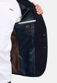 Granatowa marynarka z teksturowanej tkaniny, z widoczną gładką podszewką w ciemnym brązie i niebieskim kolorze. Elementy z etykietą „JEFF” oraz detalem kieszeni.