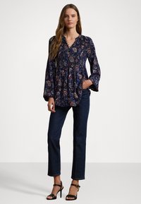 Mörkblå långärmad blus med blommönster, v-ringning, smockad midja och trumpetärm, tillsammans med mörka jeanstights och svarta klackar med remmar.