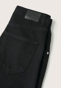 Jean en denim noir plié avec une poche arrière visible, une boucle de ceinture et une étiquette de marque en cuir noir sur la ceinture.