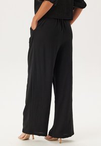 Pantalons larges noirs en tissu doux, dotés d'une ceinture élastique et de poches latérales. Texture lisse avec une coupe décontractée.