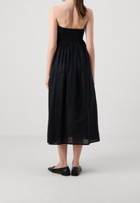 Robe longue noire sans bretelles avec un corsage smocké, une jupe fluide, et des chaussures plates noires à motif texturé. Design simple et élégant.