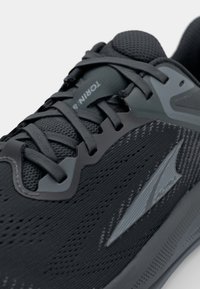 Svart sportskor med ett strukturerat mesh-övre, förstärkta plastdetaljer, platta snören och en märkeslogotyp på sidan.