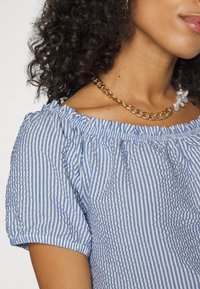 Blusa off-shoulder de riscas azuis e brancas com decote franzido e mangas curtas bufantes, combinada com um colar de corrente dourada.