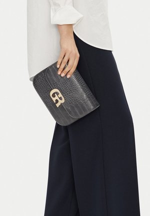 Hand holding small black crocodile-textured clutch bag with gold buckle, wearing white blouse and dark wide-leg pants.