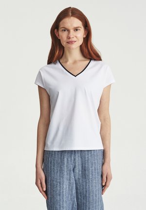 Mujer con cabello largo y rojo, vestida con una camiseta blanca de cuello en V con ribete negro y pantalones azules con rayas, de pie frente a un fondo liso.