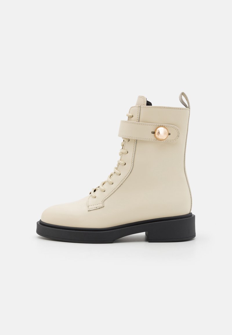Beige snørebånd ankle boot med rund tå, kontrasterende sort såle, og en rem med metallisk knapp. Glatt lærtekstur.