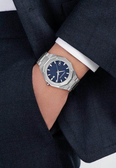 Reloj de acero inoxidable plateado con caja octogonal, esfera azul texturizada, marcadores de hora blancos y pantalla de fecha. Llevado sobre una tela de traje oscura.