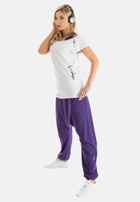 White short-sleeve top with black text and purple loose-fitting pants. Model wearing black headphones and white sneakers, posed standing.