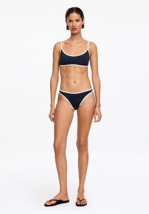 Femme debout, face à l’avant, portant un bikini noir avec un liseré blanc et des tongs noires sur fond blanc.