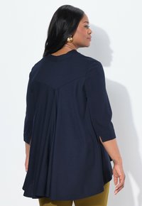 Tummansininen tunika, jossa on ¾-hihat, pyöreä pääntie ja laskostettu selkädesign. Pehmeä kangas, valuva siluetti ja sivuhalkiot.