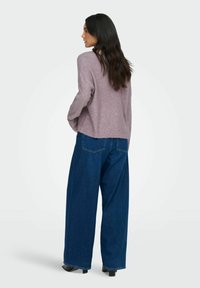 Mujer con cabello largo y oscuro lleva un suéter de color lavanda con motas, jeans de pierna ancha azul y zapatos de tacón negros, de lado frente a un fondo blanco.
