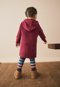 Burgunderfarbendes Strick-Hoodiekleid in mittellanger Länge mit Kapuze. Blaue und mehrfarbige gestreifte Strumpfhosen. Braune Stiefeletten. Schlichter, strukturierter Hintergrund.