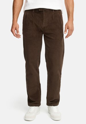 Mann trägt dunkelbraune Cordhosen und weiße Sneakers und steht vor einem weißen Hintergrund.