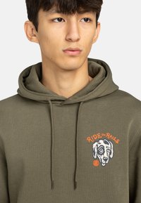 Sweatshirt vert olive avec une grande capuche, des cordons de serrage et un graphique sur le devant représentant le visage d'un chien. Le texte indique "RIDE the RAILS" en orange.