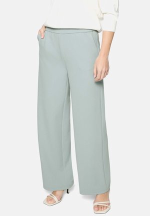 Femme portant un pantalon large bleu clair avec poches latérales et des sandales blanches à talons ouvertes, debout avec une main dans la poche.