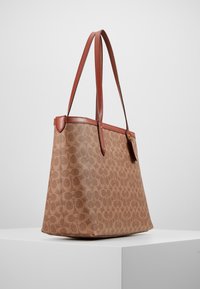 Sac cabas à motifs marron avec garniture en cuir et longues poignées, présenté sur un piédestal blanc sur fond uni.