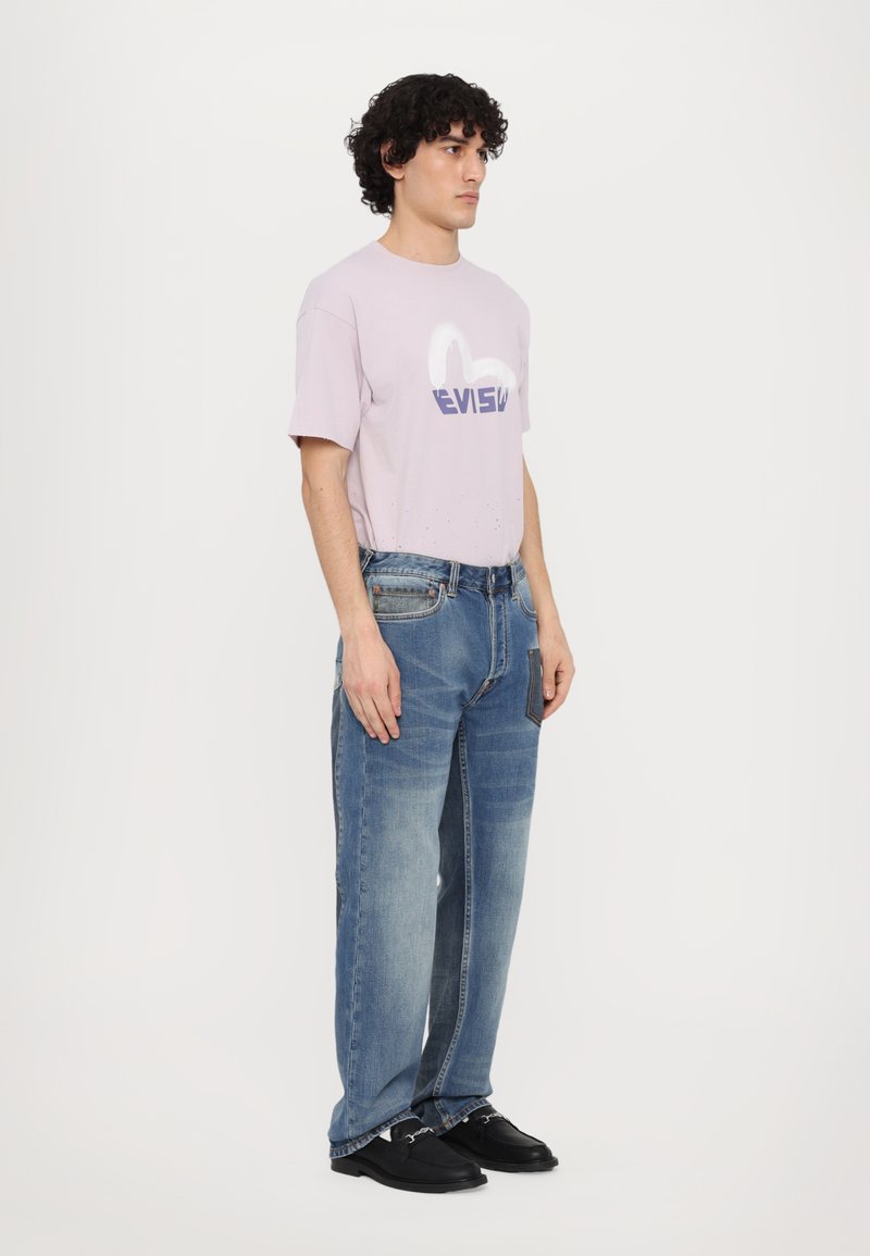 T-shirt en coton violet clair avec un logo blanc et bleu, associé à un jean bleu en denim à coupe ample et avec des poches, porté avec des chaussures noires.