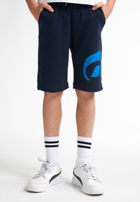 Námořnické modré sportovní šortky s velkým modrým potiskem na levé nohavici. Vyrobené z měkké tkaniny, s kapsami a volným střihem. Kombinované s bílými teniskami.