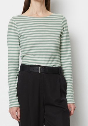 Femme portant une chemise à rayures vertes et blanches à manches longues rentrée dans un pantalon noir taille haute avec une ceinture noire.