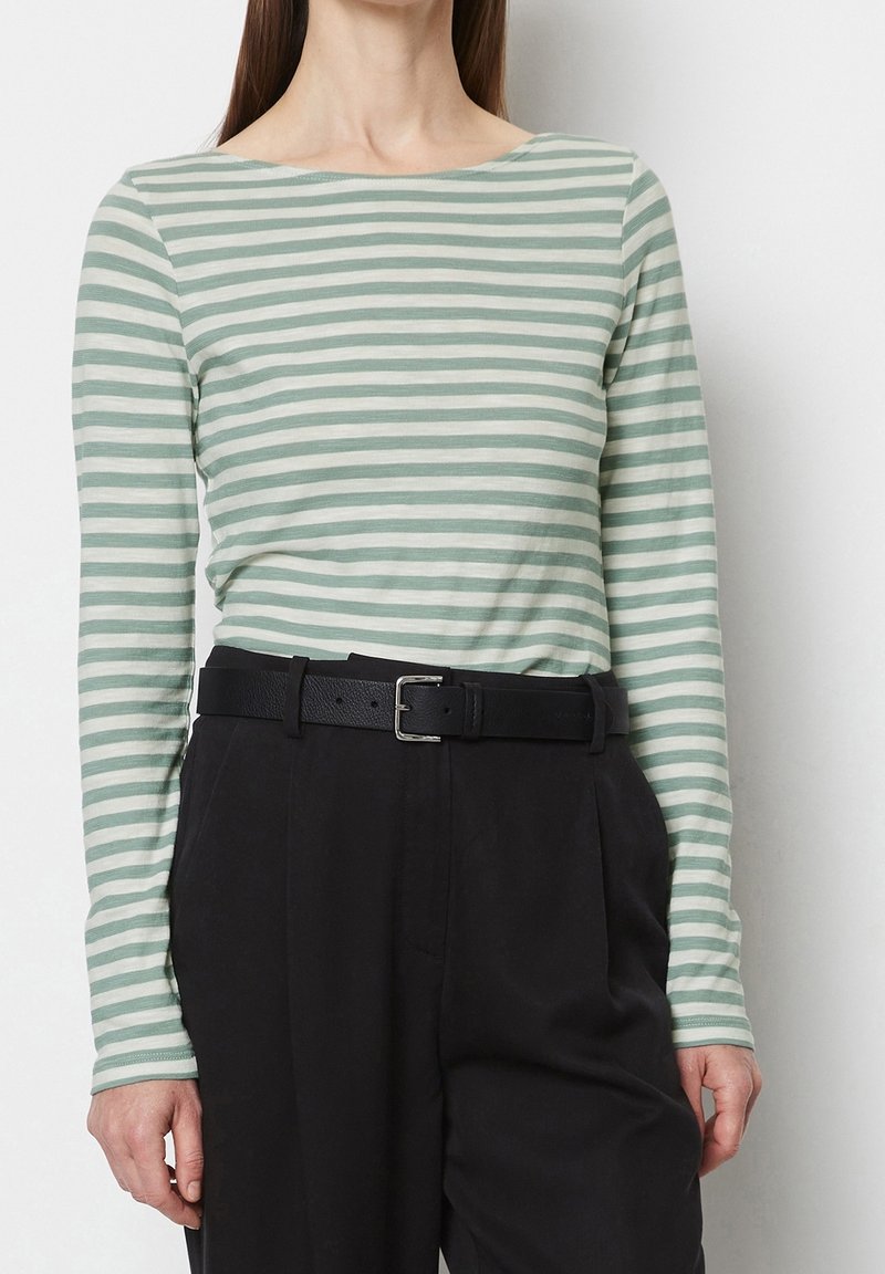 Frau in einem grün-weißen gestreiften Langarmshirt, das in eine hochgeschnittene schwarze Hose mit einem schwarzen Gürtel gesteckt ist.