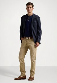 Ανδρικό ντύσιμο που περιλαμβάνει σκούρο σακάκι με υφή, μπλε πουκάμισο, μπεζ chino παντελόνι και καφέ παπούτσια. Σημαντικές λεπτομέρειες είναι η μαντηλοθήκη στο πέτο και η ζώνη.