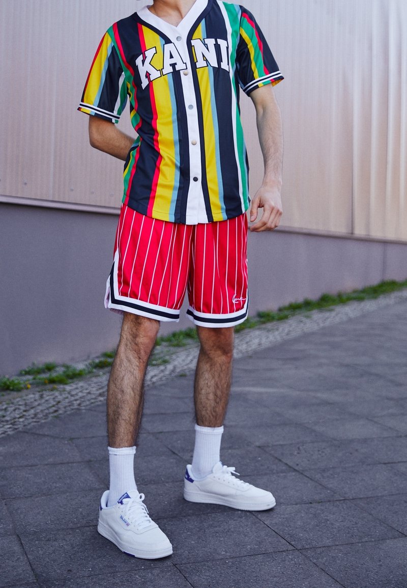 Personne portant un t-shirt rayé multicolore "KANI", un short rayé rouge et blanc, des chaussettes blanches et des baskets blanches, debout sur un trottoir à l'extérieur.