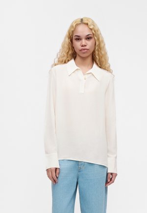 Un mannequin aux longs cheveux blonds se tient face à l’objectif, vêtu d’un chemisier blanc à col et d’un jean bleu clair, sur un fond uni.