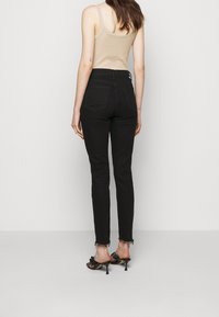 Kvinna som bär högmidjade svarta jeans med fransade fållar, beige ärmlös topp och svarta klacksandaler, står mot en enkel ljus bakgrund.