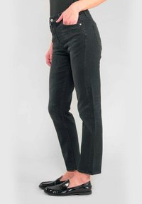 Kvinna som bär svarta rakbenade jeans och svarta lackskinnsloafers, stående mot en ljus bakgrund.